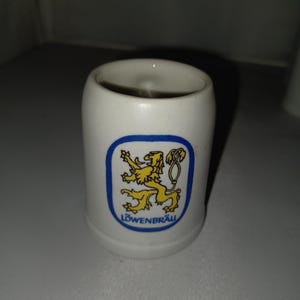 May include: White ceramic shot glass with a blue and yellow logo of a lion and the word "L&Ouml;WENBR&Auml;U". The shot glass is cylindrical and has a smooth, glossy finish. The logo is centered on the front of the glass.