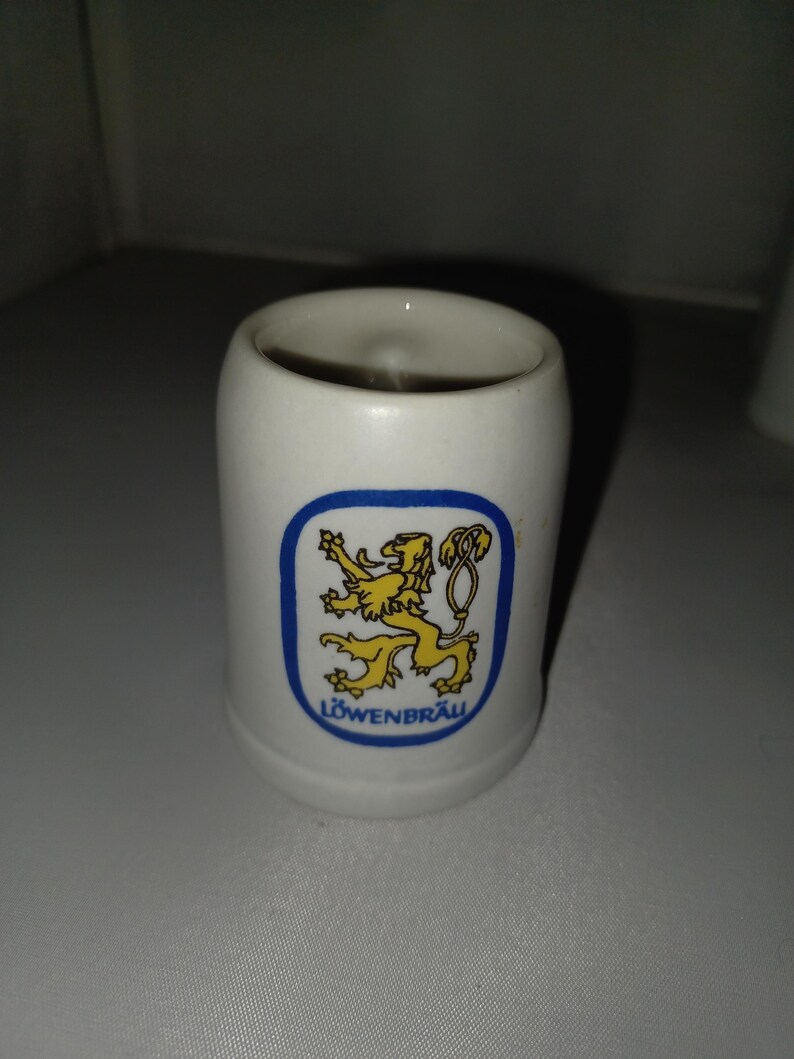 May include: White ceramic shot glass with a blue and yellow logo of a lion and the word "L&Ouml;WENBR&Auml;U". The shot glass is cylindrical and has a smooth, glossy finish. The logo is centered on the front of the glass.