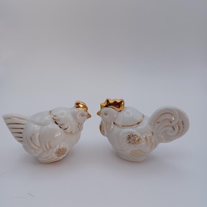 May include: Two white ceramic rooster figurines with gold accents. One rooster is standing, the other is sitting. The figurines have gold detailing on the comb, wings, and tail. The background is plain white.