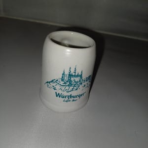May include: A white ceramic shot glass with a teal illustration of a castle and the words "W&uuml;rzburger Light Beer". The glass has a cylindrical shape and a slightly flared top.