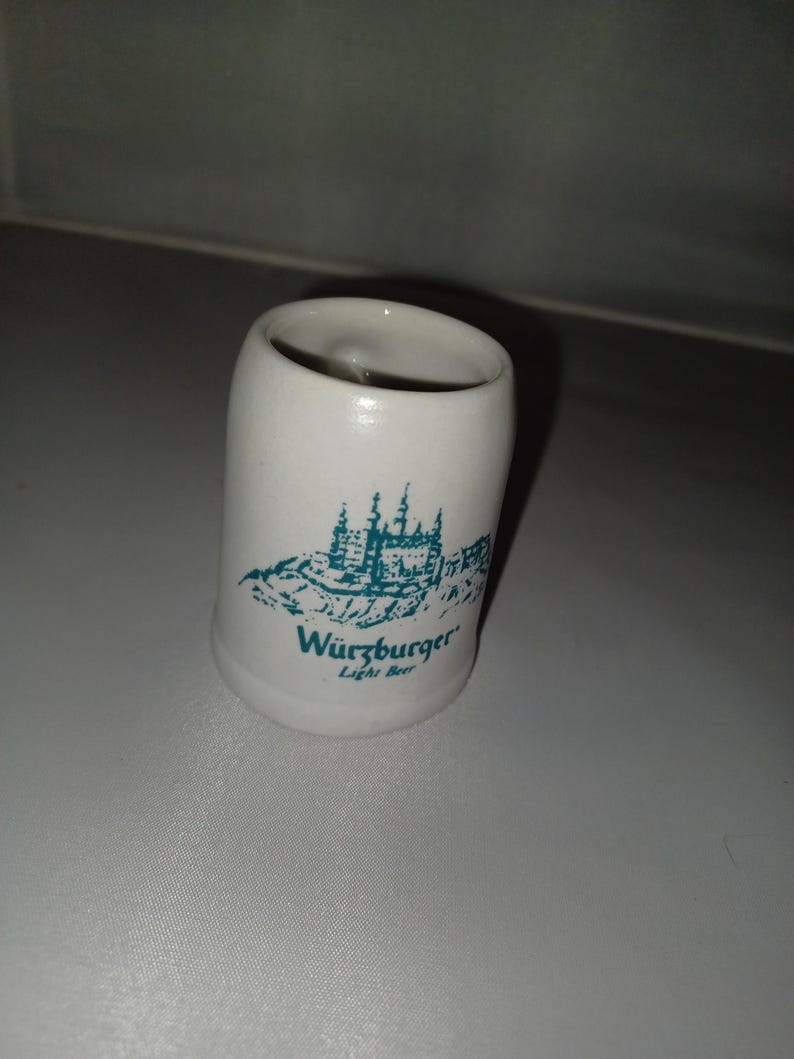 May include: A white ceramic shot glass with a teal illustration of a castle and the words "W&uuml;rzburger Light Beer". The glass has a cylindrical shape and a slightly flared top.