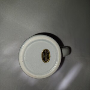 May include: A white ceramic mug with a handle, viewed from the bottom. A gold label with the text "Ceramarte Made in Brazil" is affixed to the base. The mug has a light blue interior.