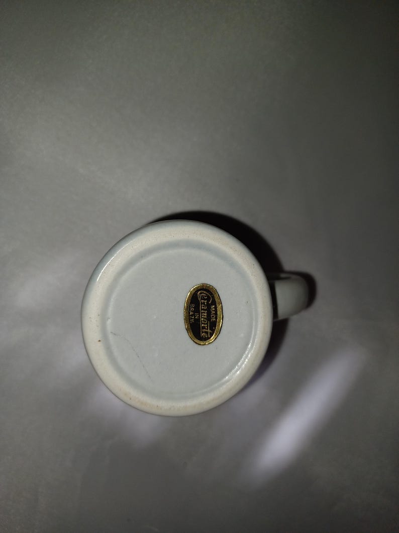 May include: A white ceramic mug with a handle, viewed from the bottom. A gold label with the text "Ceramarte Made in Brazil" is affixed to the base. The mug has a light blue interior.