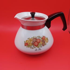 May include: A white teapot with a black handle and a silver-toned lid featuring a black knob. The body of the teapot is decorated with a colorful botanical illustration of fruits and flowers, with the text Le Thé printed in black script below.