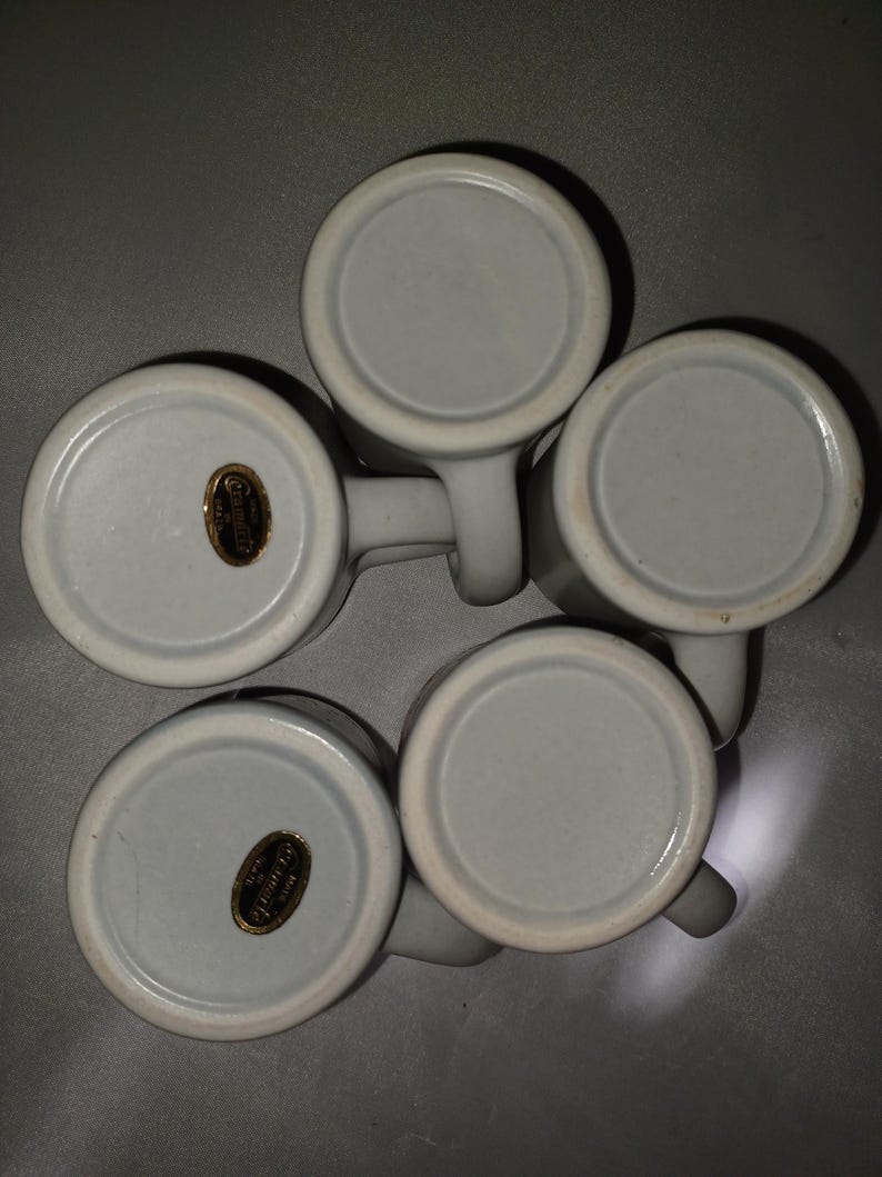 May include: Five white ceramic mugs with handles, arranged in a circular pattern. The bottoms of the mugs are visible, showing a light gray color and a gold-colored sticker. The mugs are likely for beverages.