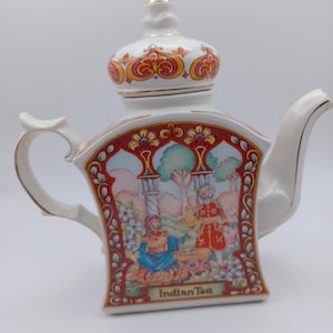 May include: White ceramic teapot with a decorative lid and handle. The teapot features a colorful illustration of an Indian scene with the words "Indian Tea" at the bottom. The design includes red, yellow, and blue accents, with gold trim.
