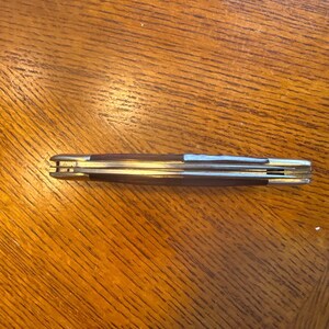 May include: A closed folding pocket knife featuring a dark brown wooden handle with polished silver-tone metal bolsters at both ends. The knife has a slim, elongated profile with visible metal liners and a spine showing the folded blade edges.