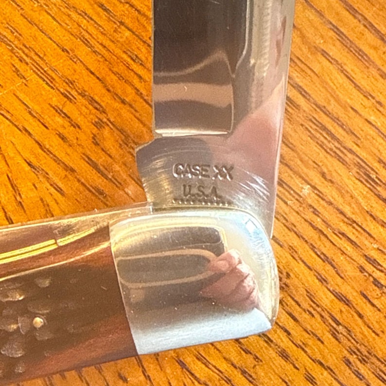 May include: A folding pocket knife with a dark brown textured handle and a polished metal bolster. The silver-colored blade features the stamped text CASE XX U.S.A. near the base, reflecting light on its smooth, metallic surface.