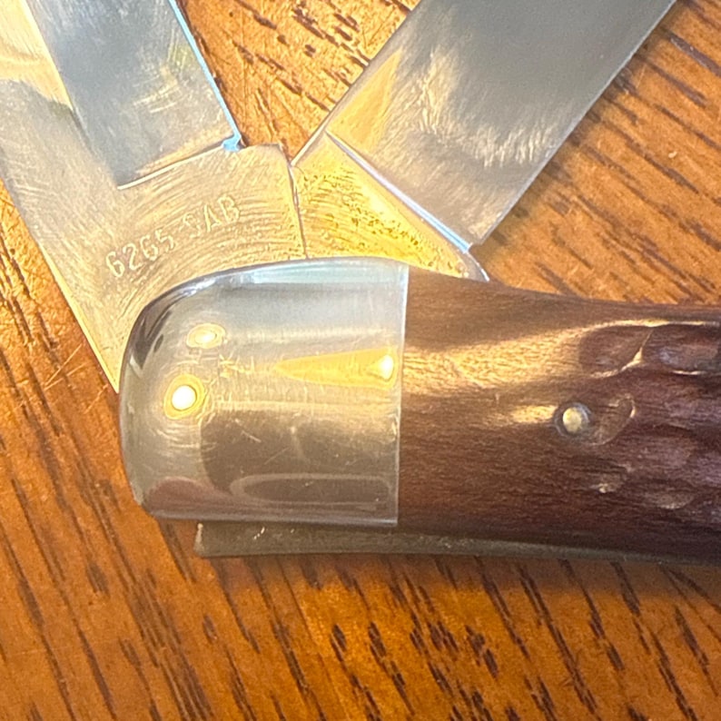 May include: A folding pocket knife with a dark brown textured handle and a polished metal bolster. The metal blade is partially visible and features the stamped text 6265 SAB. The knife is shown in an open position with two distinct blade sections.