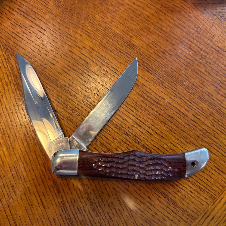 May include: A folding pocket knife with two silver-colored metal blades extended at an angle. The handle features a dark brown, textured wood-grain finish with polished silver-colored metal bolsters at both ends and a small pin near the base.
