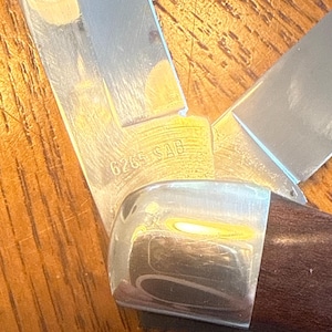 May include: A folding pocket knife with a dark brown wooden handle and a polished metal bolster. The silver-colored metal blade features the engraved text 6265 SAB near the base, showing a sharp edge and a metallic finish with visible reflections.