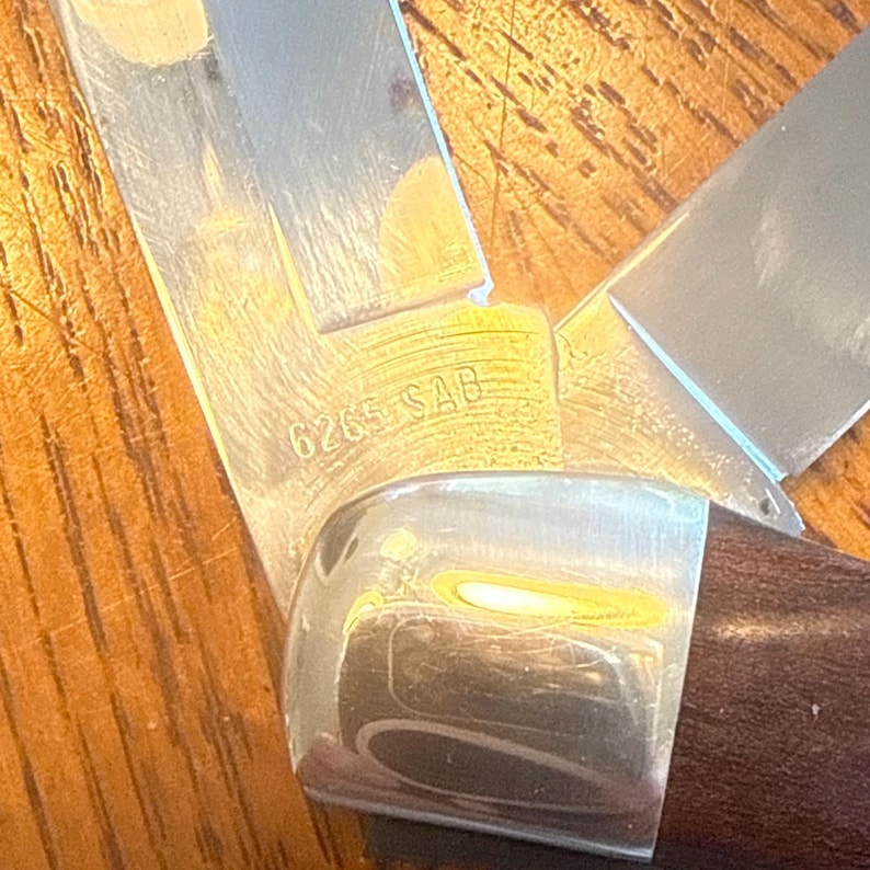 May include: A folding pocket knife with a dark brown wooden handle and a polished metal bolster. The silver-colored metal blade features the engraved text 6265 SAB near the base, showing a sharp edge and a metallic finish with visible reflections.