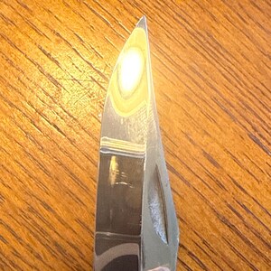 May include: A sharp, pointed metal knife blade with a polished, reflective surface. The blade features a curved spine that tapers to a fine tip, with a distinct nail nick groove visible along the side of the metallic steel construction.