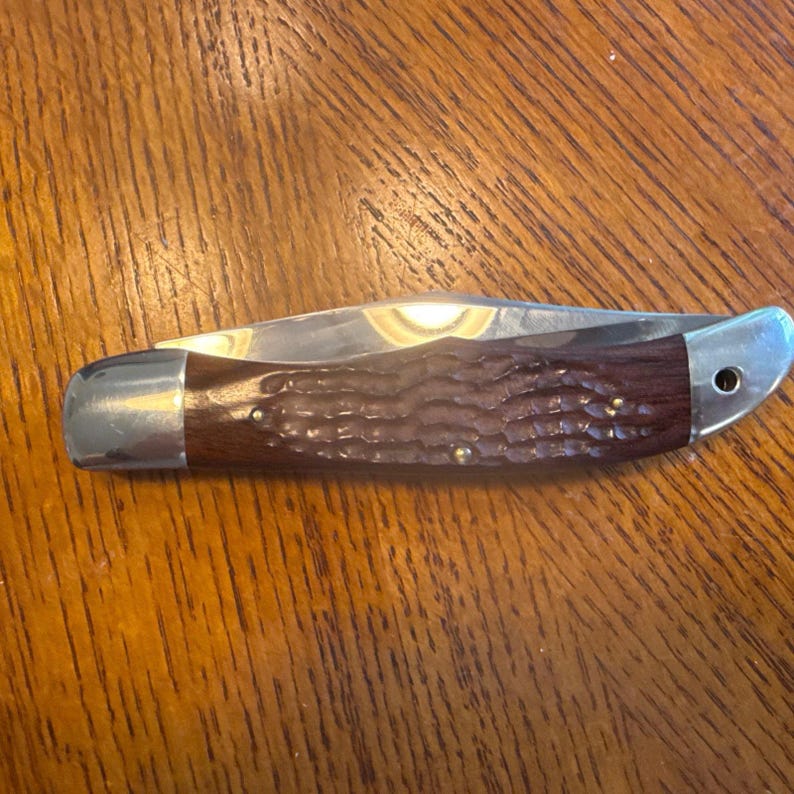 May include: A folding pocket knife with a dark brown, textured wooden handle and polished silver-tone metal bolsters at both ends. The blade is partially visible, tucked into the handle, and features a small hole near the rear bolster.