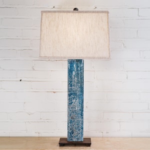 May include: A tall, rectangular table lamp with a textured blue base and a rectangular beige lampshade. The lamp is set on a dark brown base and stands against a white brick wall.