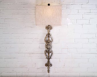 May include: A wall-mounted lamp with a rectangular beige lampshade. The lamp has an ornate, decorative metal base with scrollwork and floral designs. The lamp is mounted on a white brick wall.