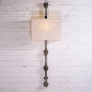 May include: A wall-mounted lamp with a rectangular, off-white lampshade. The lamp is supported by a decorative, dark metal rod with ornate detailing. The lamp is mounted on a white brick wall.