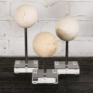 May include: Three decorative spheres with a mottled, off-white finish. Each sphere is mounted on a dark metal rod and set on a clear acrylic base. The spheres vary slightly in size and are arranged in a group.