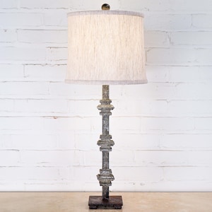 May include: A tall, ornate lamp with a textured, off-white lampshade. The lamp's base and column are a weathered gray with decorative details. The lamp stands on a square base and is set against a white brick wall.