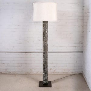 May include: A tall floor lamp with a textured, dark gray column and a square base. The lamp has a white, cylindrical lampshade. The lamp is positioned in front of a white brick wall.