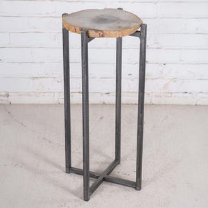 May include: A tall, modern plant stand with a round, light-colored wood top. The top is supported by a dark gray, metal frame with four legs and an X-shaped base. The stand is designed for indoor use.