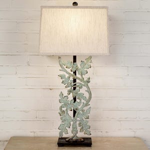 May include: A decorative table lamp with a light beige rectangular lampshade. The lamp base features a detailed, openwork design of leaves and vines in a distressed, pale green finish. The lamp is set against a white brick wall.