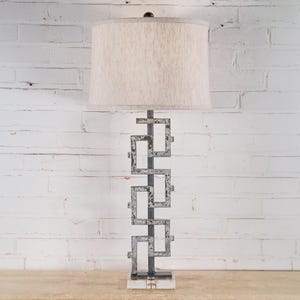 May include: A tall, decorative table lamp with a rectangular base and a geometric metal frame. The lamp has a beige fabric shade and a dark metal finial. The lamp is approximately 30 inches tall.