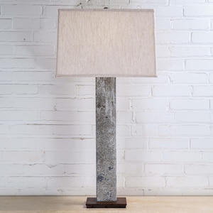 May include: A tall floor lamp with a rectangular beige lampshade. The lamp's base is a dark brown, and the column is a textured gray. The background is a white brick wall.