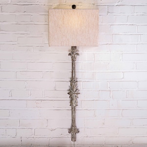 May include: A wall-mounted lamp with a rectangular beige lampshade. The lamp has a decorative, ornate metal base with floral and leaf designs. The lamp is mounted on a white brick wall.