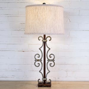May include: A decorative table lamp with a beige fabric shade. The lamp features a dark brown metal base with ornate, scroll-like accents in a silver finish. The lamp is set against a white brick wall.