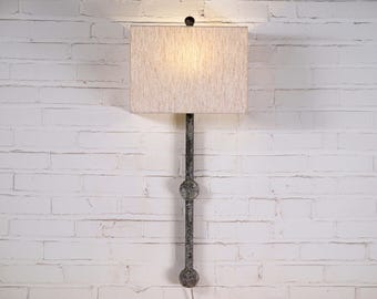 May include: A wall-mounted lamp with a rectangular, off-white lampshade. The lamp has a dark, textured metal stem with decorative circular elements. The lamp is mounted on a white brick wall.