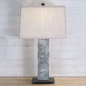 May include: A rectangular table lamp with a gray, textured base and a beige rectangular lampshade. The lamp is set against a white brick wall. The lamp base appears to be made of stone or a stone-like material.
