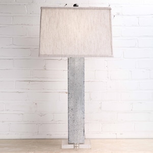 May include: A tall, modern floor lamp with a rectangular lampshade. The lampshade is a light beige color. The lamp's base is a textured, gray concrete column. The lamp is set on a clear acrylic base. The background is a white brick wall.