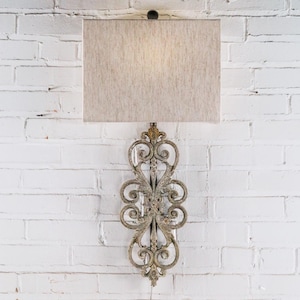 May include: A wall-mounted lamp with a rectangular beige lampshade. The lamp features an ornate, scrollwork design in a distressed gold finish. The lamp is mounted on a white brick wall.