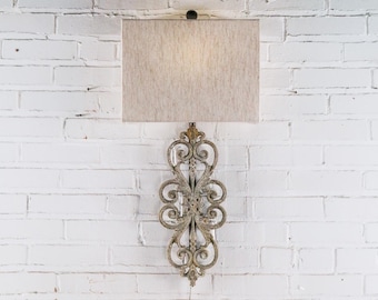 May include: A wall-mounted lamp with a rectangular beige lampshade. The lamp features an ornate, scrollwork design in a distressed gold finish. The lamp is mounted on a white brick wall.