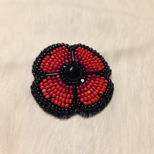 May include: A beaded flower brooch with a black outline and a black central bead. The flower petals are red, with darker red petals in the centre. The brooch is set against a white, fluffy background. A handcrafted accessory for a touch of floral elegance.