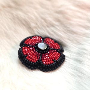 May include: A beaded poppy flower brooch featuring red and black beads. The flower has a silver metal centre and is set against a white, fur-like background. The poppy design is a classic floral motif.