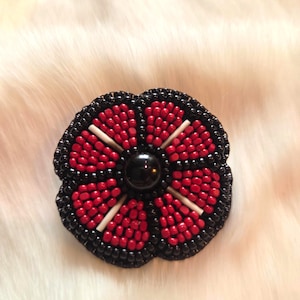 Puede incluir: Un broche de flores con cuentas con un borde negro, pétalos rojos y una cuenta negra en el centro. Los pétalos tienen detalles blancos. El broche está sobre un fondo blanco y esponjoso.