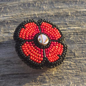 Pode incluir: Broche de flores artesanal com contas vermelhas e pretas. As pétalas são vermelhas, a borda externa é preta e o centro tem uma conta iridescente. Este broche floral é um acessório único.