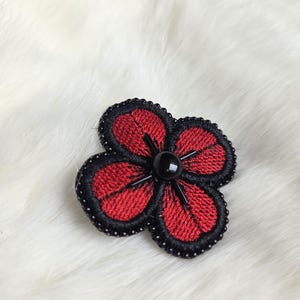 Peut inclure: Une broche fleur brodée à trois pétales. Les pétales rouges sont bordés de points noirs et de perles noires. Une perle noire est au centre de la fleur.