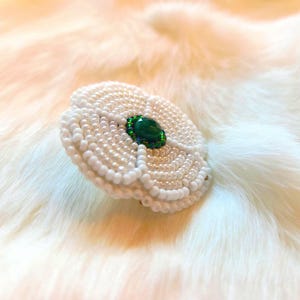 May include: A white beaded flower brooch with a central green gemstone. The brooch is crafted from small, round white beads, forming a layered petal design. The gemstone is a deep green, encircled by smaller green beads.