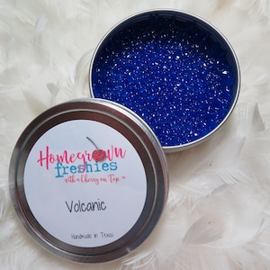 May include: A silver tin with a blue lid filled with blue gel beads. The tin has a white label with the text "Homegrown Freshies with a Cherry on Top." and "Volcanic" in pink lettering. The label also says "Handmade in Texas".