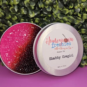 May include: A silver tin with a pink and black glittery substance inside. The tin has a label that reads "Homegrown Freshies with a cherry on top" and "Shabby Cowgirl". The label also says "Freshie Tin" and "Made in Texas".