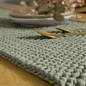 May include: A sage green knitted table runner with a textured surface. The runner is made of thick, chunky yarn and has a subtle sheen.