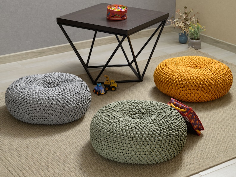 May include: Three round knitted poufs in gray, green, and yellow. The poufs are sitting on a beige rug in front of a black metal coffee table.