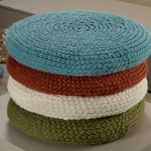 May include: Four round knitted cushions stacked on top of each other. The top cushion is teal blue, the next is rust orange, the next is off-white, and the bottom cushion is olive green.