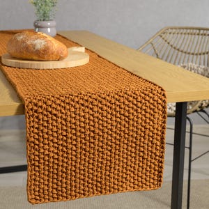 May include: A brown knitted table runner with a textured pattern. The runner is placed on a wooden table with a loaf of bread on a wooden chopping board.