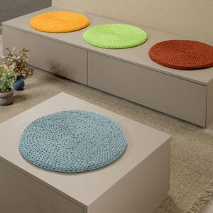 May include: Four round crocheted cushions in different colours, orange, green, red, and blue, are placed on a light grey bench and a light grey coffee table. The bench and table are on a beige rug with fringe.