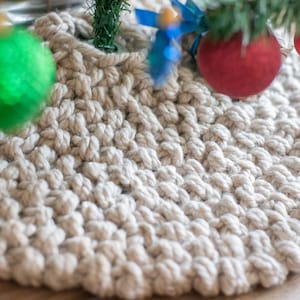 Op de afbeelding: Een crèmekleurige gehaakte boomrok met een groene, rode en blauwe kerstversiering erop.