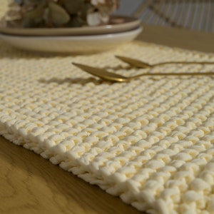 Könnte beinhalten: Nahaufnahme eines cremefarbenen gestrickten Tischläufers mit strukturierter Oberfläche. Der Läufer liegt auf einem Holztisch.